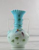 Opalhvid vase 
med blå 
omfangsglas. 
Bemalet med 
farvestrålende 
fugl. Vasen har 
to hanke.
H. 20,5 ...