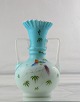 Opalhvid vase 
med blå 
omfangsglas. 
Bemalet med 
farvestrålende 
fugl. Vasen har 
to hanke.
H. 20,5 ...