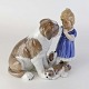 Figur i 
porcelæn med 
motiv af lille 
pige som 
klapper en 
Sankt Bernhard 
tæve, mens 
tævens hvalpe 
...